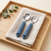 Cubkins blue olive silicone-handle 304 steel toddler spoon and fork on a linen napkin on a wooden tray