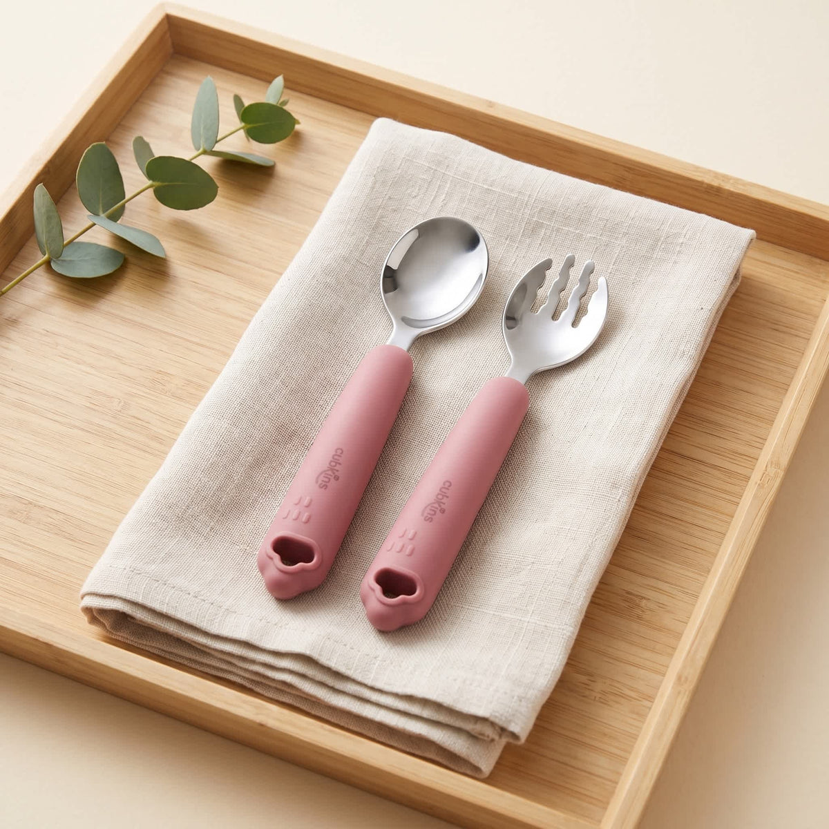 Cubkins raspberry silicone-handle 304 steel toddler spoon and fork on a linen napkin on a wooden tray