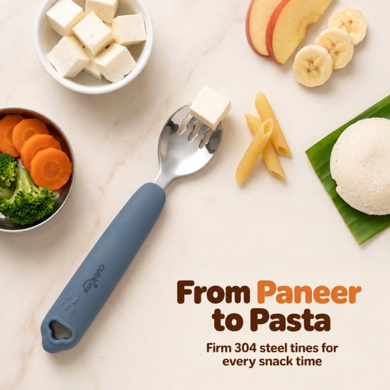 Cubkins blue olive 304 steel toddler fork with a paneer cube on the tines, surrounded by paneer, idli on banana leaf, carrots, broccoli, pasta and fruit