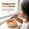 Indian toddler holding the Cubkins blue olive steel fork in a palmar grasp and bringing a carrot coin to their mouth