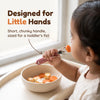 Indian toddler holding the Cubkins raspberry steel fork in a palmar grasp and bringing a carrot coin to their mouth