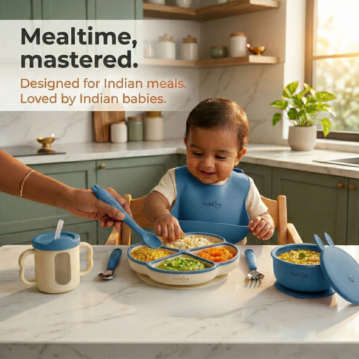 Baby wearing a blue olive Cubkins silicone bib eating from a divided suction plate at mealtime, with suction bowl, sensory straw training cup with transparent window, steel spoon and fork alongside