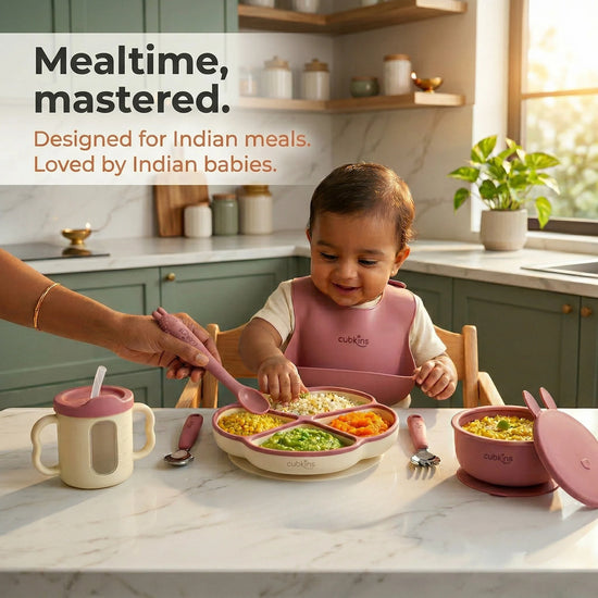 Baby wearing a raspberry Cubkins silicone bib eating from a divided suction plate at mealtime, with suction bowl, sensory straw training cup with transparent window, steel spoon and fork alongside