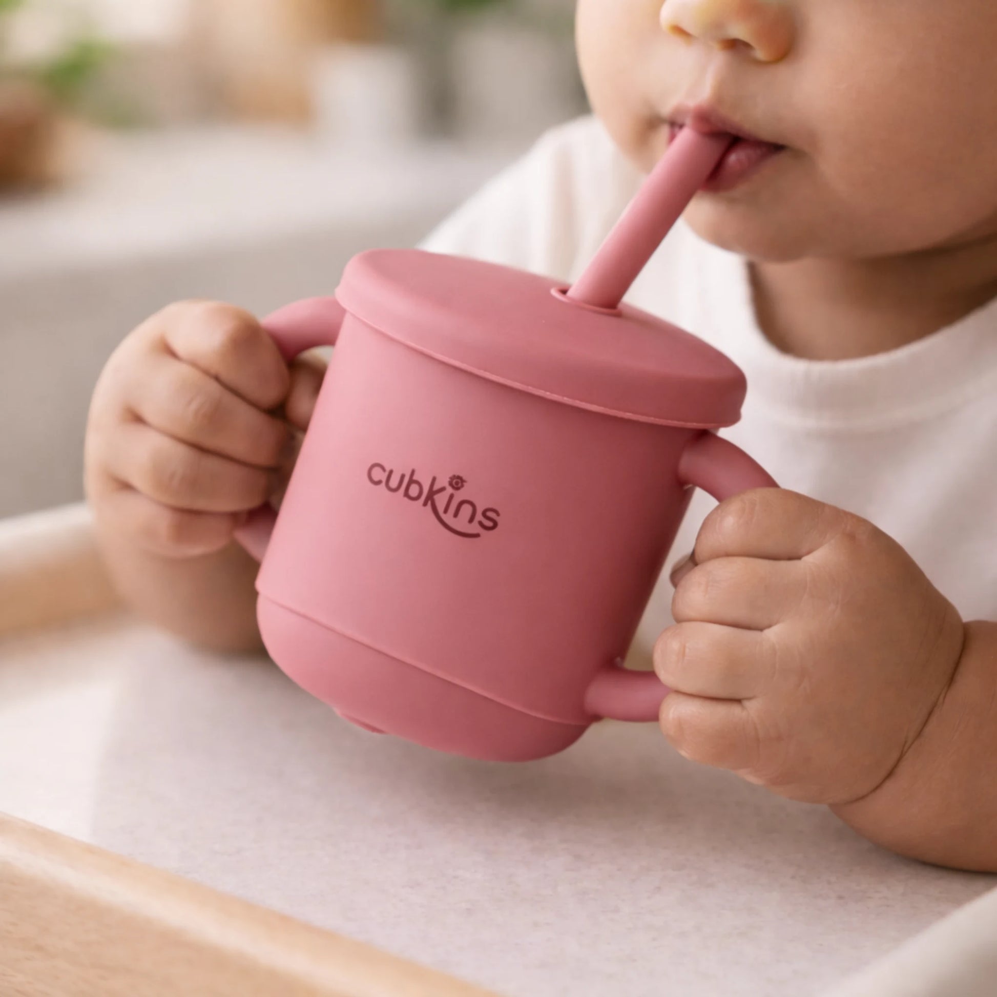 Indian baby holding and drinking independently from the Cubkins silicone straw cup in Raspberry, seated in a high chair