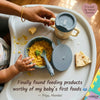 Overhead view of a baby's high chair tray with Cubkins blue suction bowl filled with khichdi, roti piece, and training cup — with Indian mom's hand and gold bangle in frame. Quote from Priya, Mumbai.