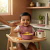 Indian baby in a high chair eating khichdi from a Cubkins raspberry silicone suction bowl with bendable spoon and training cup on the tray — made for Indian mealtimes.
