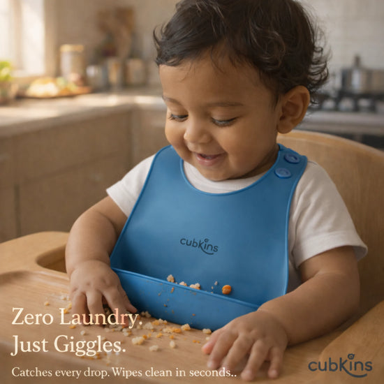 Happy toddler wearing Cubkins blue silicone bib during mealtime in kitchen, demonstrating zero laundry easy-clean design