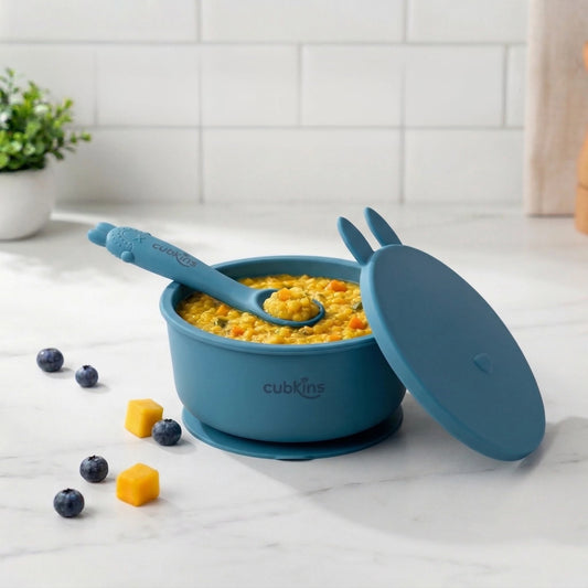 Cubkins blue silicone baby feeding bowl with suction base, bunny ear lid, and curved spoon with food on marble counter