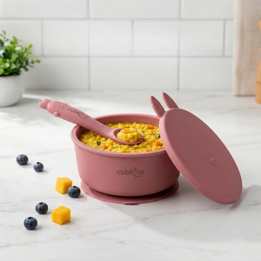 Cubkins raspberry pink silicone baby feeding bowl with suction base, bunny ear lid, and curved spoon with food on marble counter