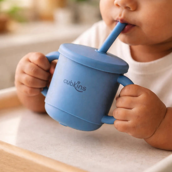 Indian baby in a high chair holding the Cubkins blue olive silicone straw cup by both handles and drinking through the straw