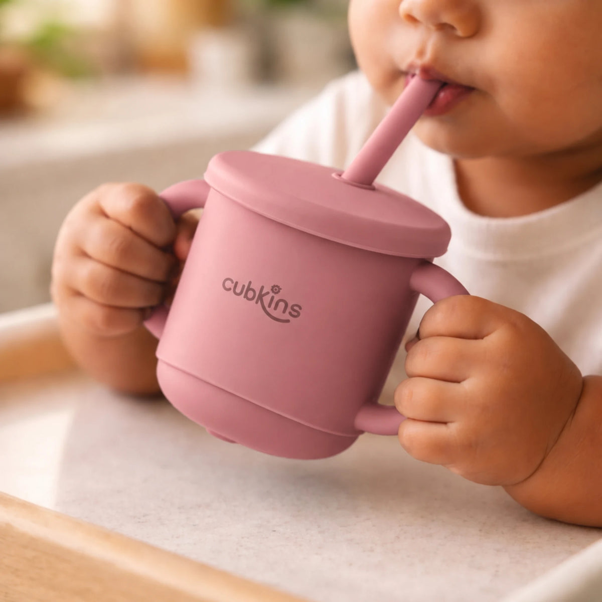 Indian baby in a high chair holding the Cubkins raspberry silicone straw cup by both handles and drinking through the straw