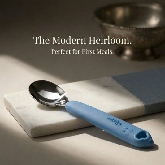 Cubkins stainless steel baby spoon with blue olive silicone handle displayed on a white marble surface