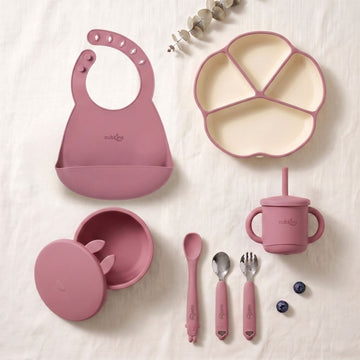 Cubkins 7-piece essential silicone baby feeding set in raspberry pink — bib, 4-compartment suction plate, suction bowl with lid, bendable spoon, steel spoon, steel fork and silicone straw cup laid out flat