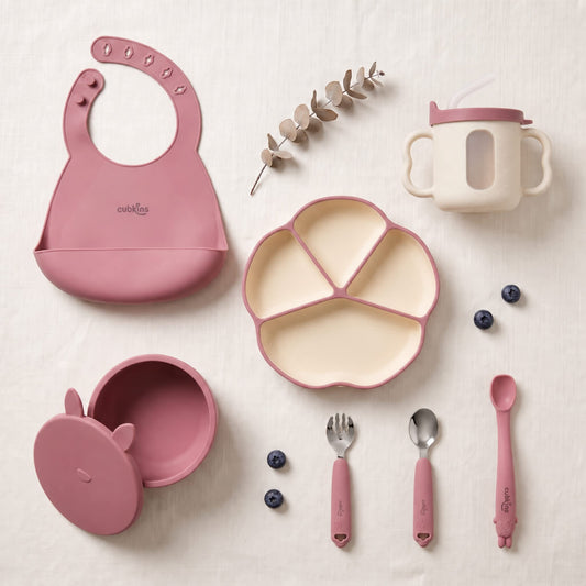 Cubkins 7-piece essential silicone baby feeding set in raspberry pink — bib, 4-compartment suction plate, suction bowl with lid, bendable spoon, steel spoon, steel fork and sensory straw training cup with transparent window laid out flat
