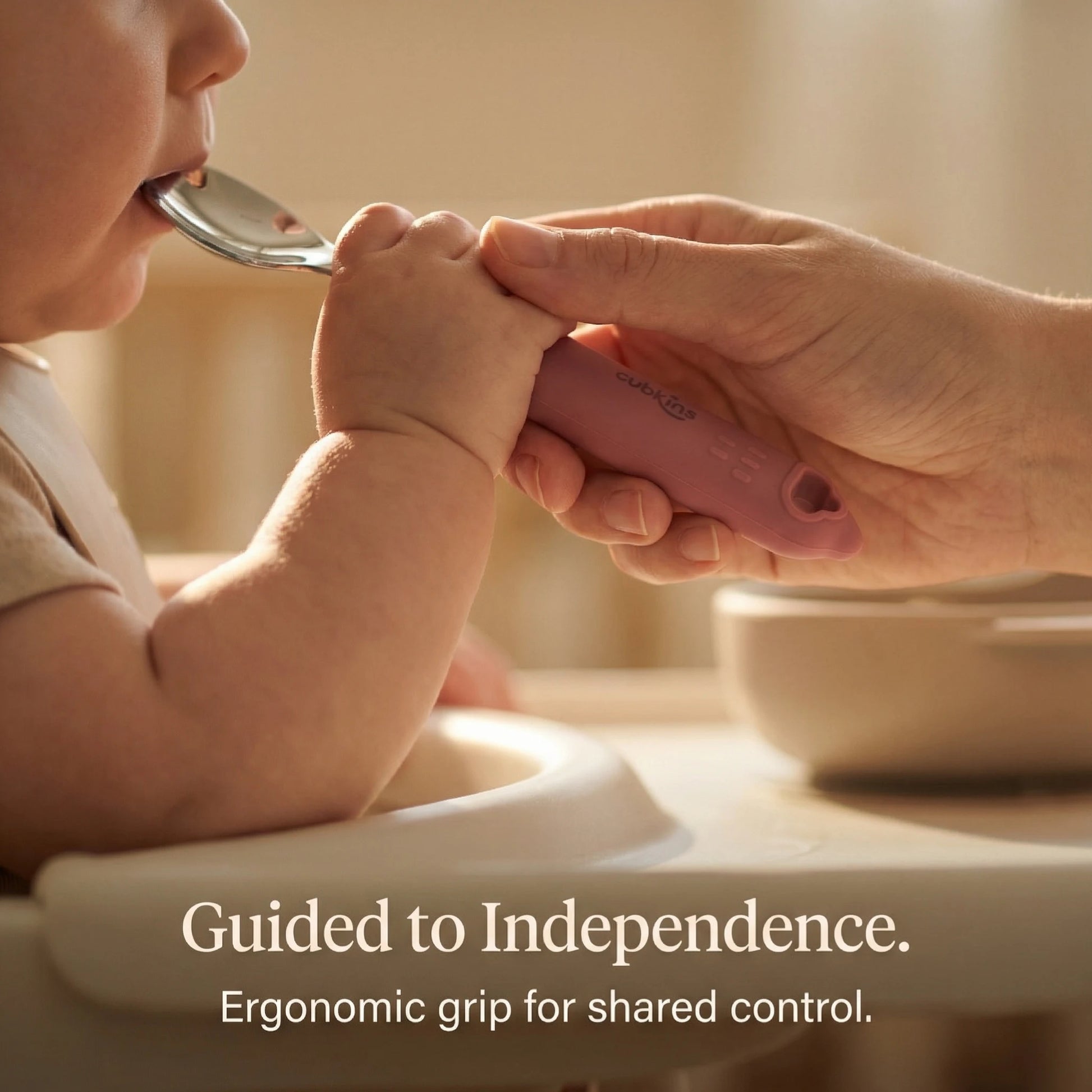 Adult hand guiding a toddler's hand around the ergonomic silicone handle of the Cubkins baby spoon