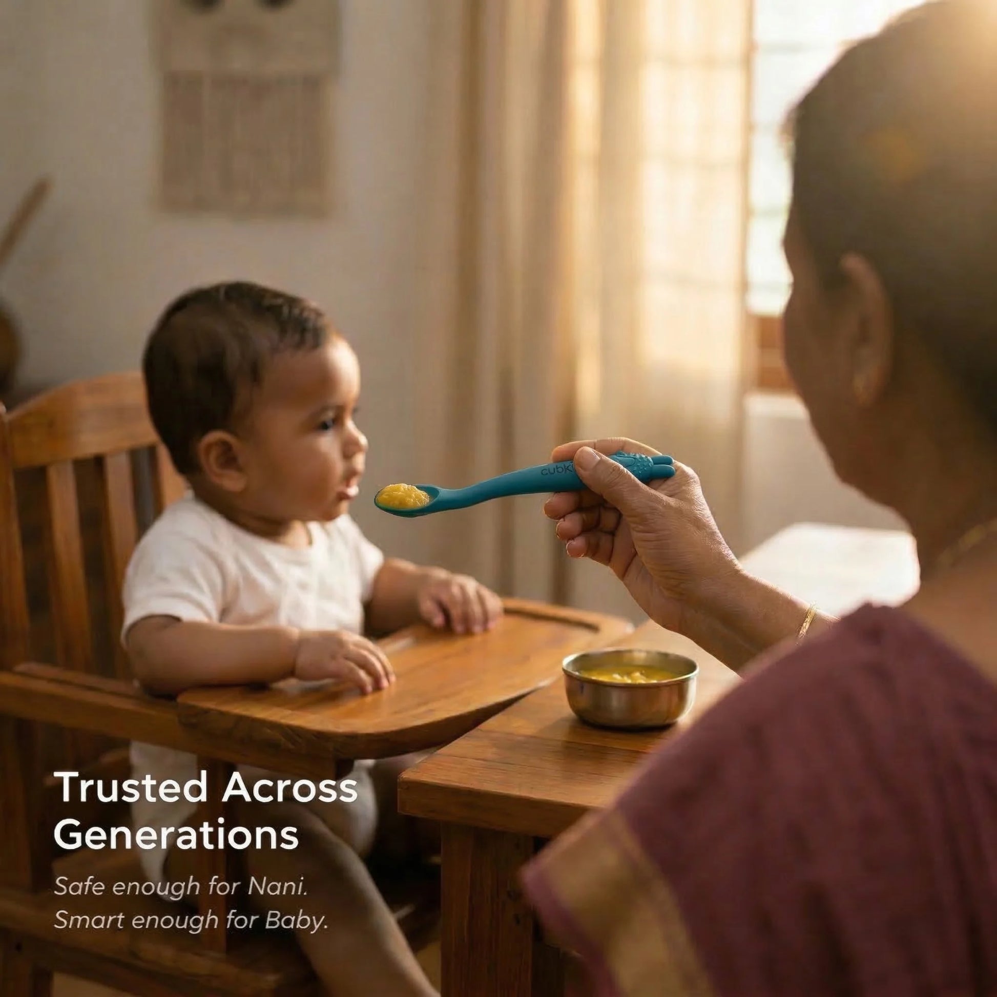 Indian grandmother (Nani) feeding a baby in a wooden high chair using the Cubkins blue silicone baby spoon from a steel bowl, in warm home setting