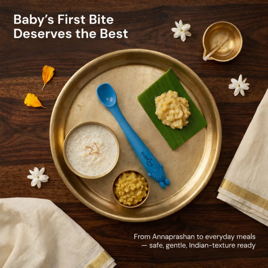Cubkins blue silicone baby spoon on a traditional brass Annaprashan thali with payasam, banana mash, and dal, decorated with marigold and jasmine flowers
