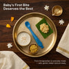 Cubkins blue silicone baby spoon on a traditional brass Annaprashan thali with payasam, banana mash, and dal, decorated with marigold and jasmine flowers