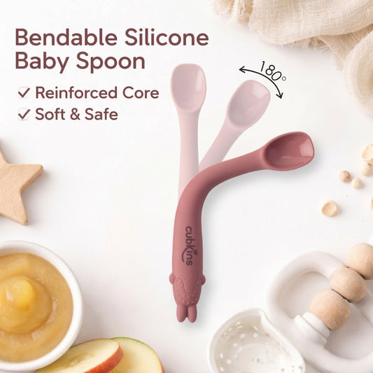 Cubkins bendable silicone baby spoon in raspberry pink bending 180 degrees, showing reinforced core and soft safe design, surrounded by baby food and wooden toys