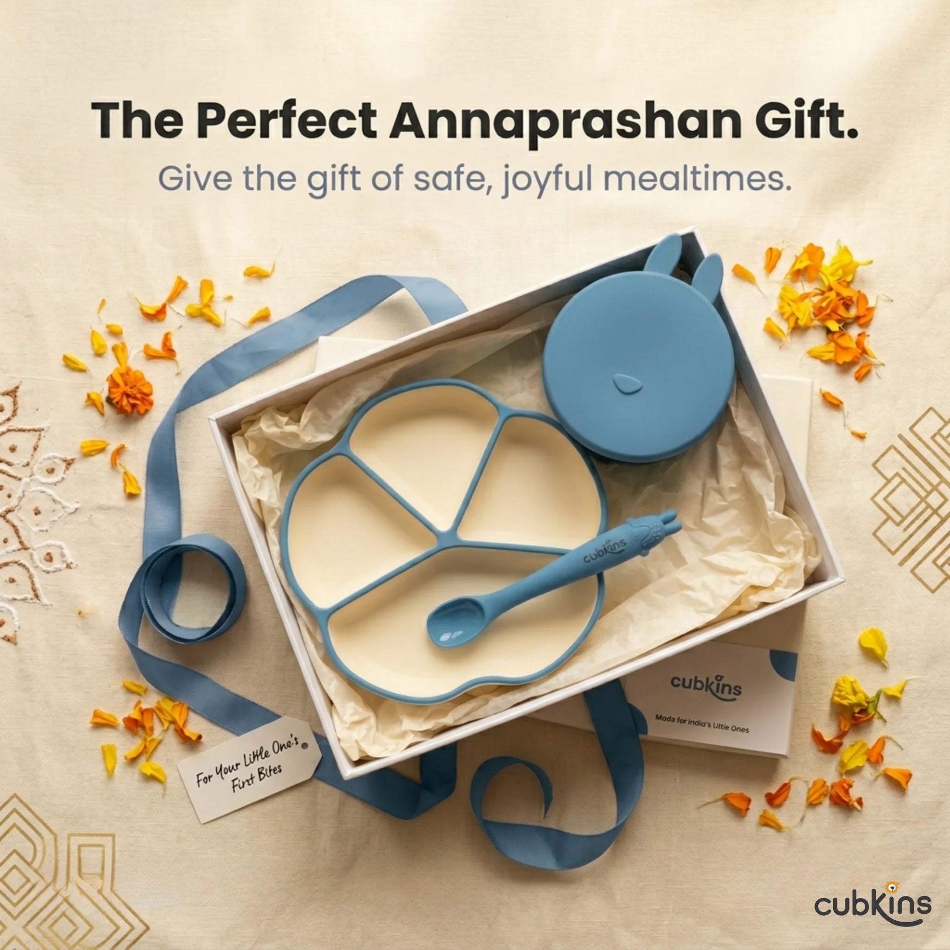 Cubkins Blue Olive plate, bowl, and bendable spoon set presented in an open gift box with tissue paper, blue satin ribbon, and scattered marigold petals on an embroidered fabric background — perfect Annaprashan or baby shower gift