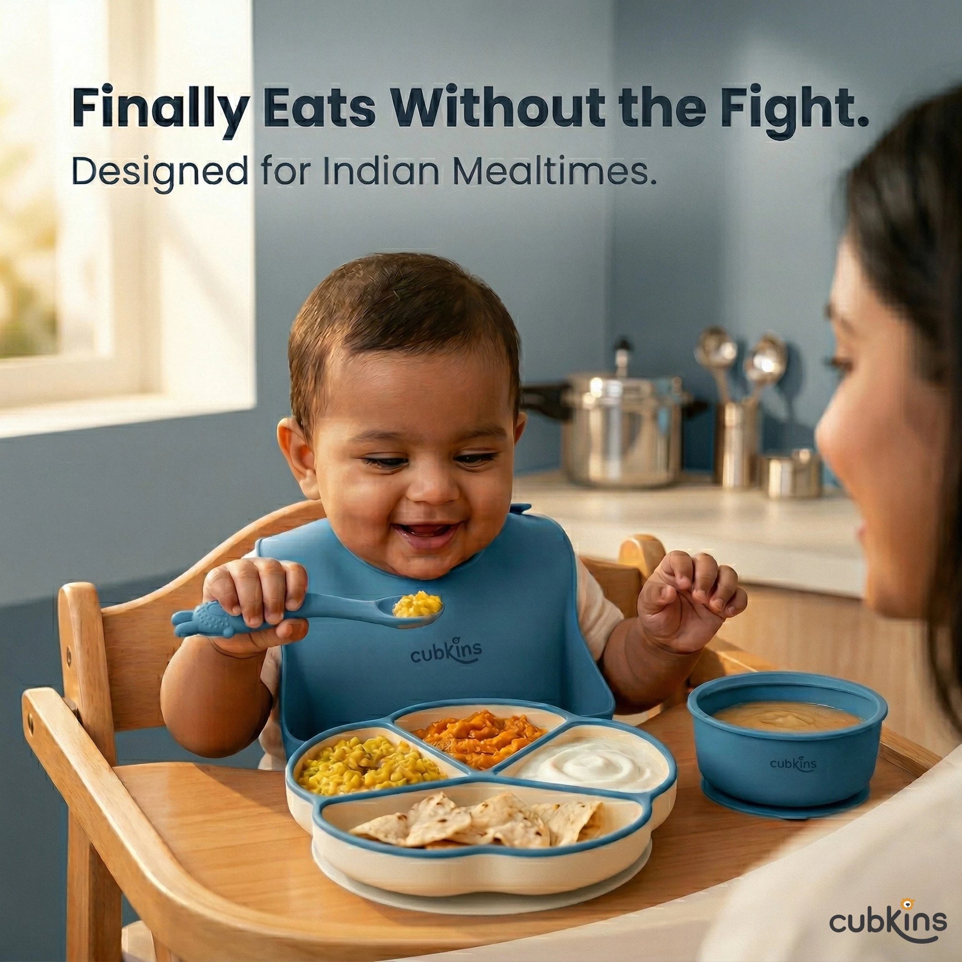 Smiling Indian baby in a highchair self-feeding with a Cubkins blue bendable spoon from a 4-compartment suction plate loaded with dal, sabzi, dahi, and roti — mother watching from the side