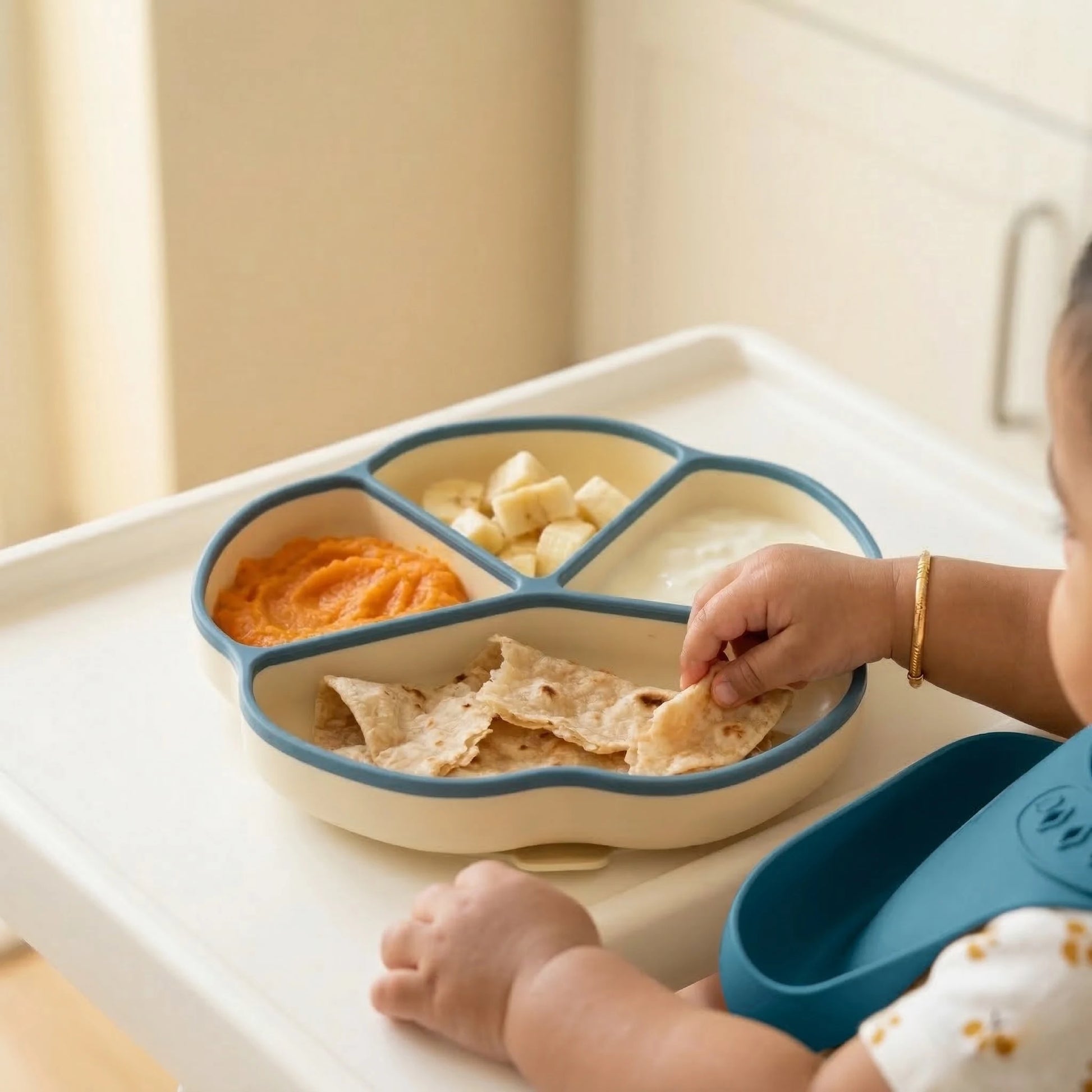 Baby self-feeding from blue Cubkins suction plate on highchair tray, picking up roti with sweet potato purée and banana cubes