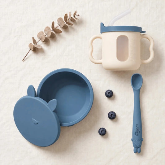 Cubkins feeding set in Blue Olive: silicone suction bowl with its bunny storage lid, sensory straw training cup and Smart-Bend bendable spoon, top-down on linen
