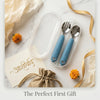 Cubkins blue toddler cutlery set in travel case styled as a gift with marigold flowers, gold ribbon, a silk pouch, and an Aashirvaad blessing card on a marble surface.