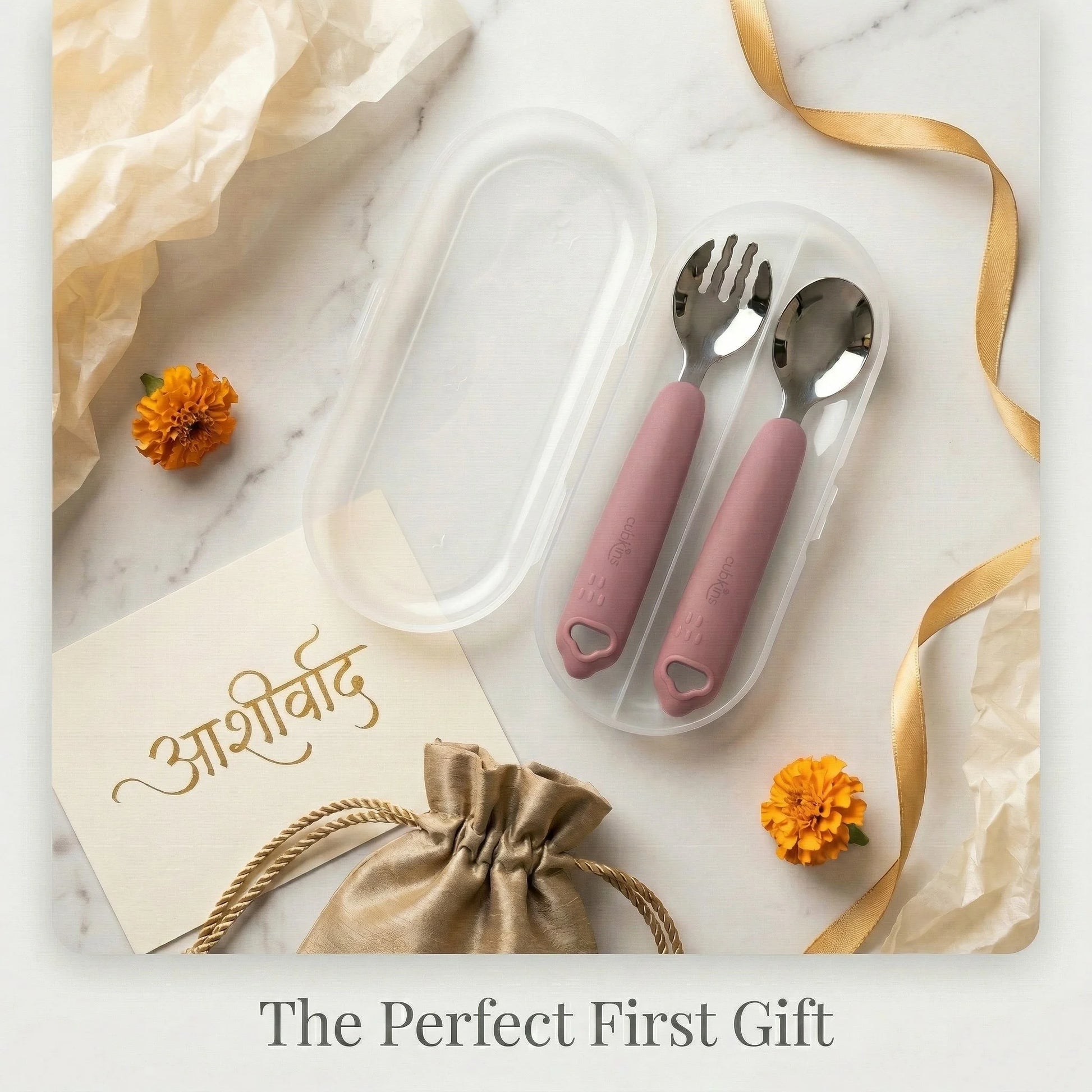 Cubkins raspberry toddler cutlery set in travel case presented as a baby gift alongside marigold flowers, gold ribbon, a satin pouch, and an Aashirvaad blessing card