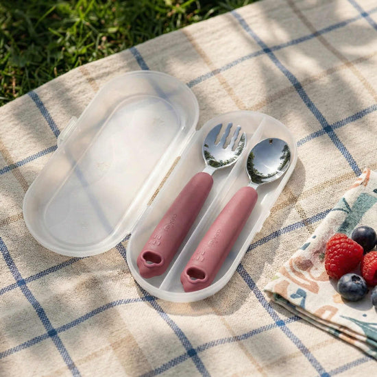 Cubkins raspberry pink toddler fork and spoon set inside an open clear travel case on a picnic blanket