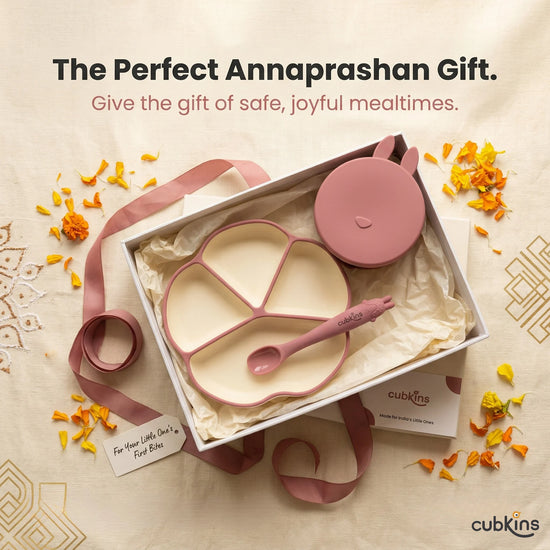 Cubkins Raspberry plate, bowl, and bendable spoon set presented in an open gift box with tissue paper, raspberry satin ribbon, and scattered marigold petals on an embroidered fabric background — perfect Annaprashan or baby shower gift