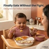 Smiling Indian baby in a highchair self-feeding with a Cubkins raspberry bendable spoon from a 4-compartment suction plate loaded with dal, sabzi, dahi, and roti — mother watching from the side