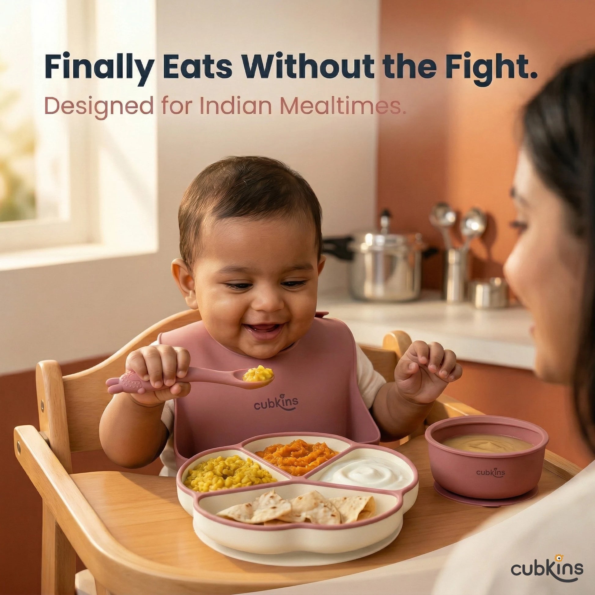 Smiling Indian baby in a highchair self-feeding with a Cubkins raspberry bendable spoon from a 4-compartment suction plate loaded with dal, sabzi, dahi, and roti — mother watching from the side