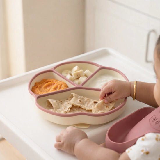 Baby self-feeding from raspberry Cubkins suction plate on highchair tray, picking up roti with sweet potato purée and banana cubes