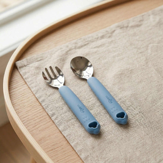 Cubkins blue silicone-handled stainless steel toddler fork and spoon set laid on a linen napkin on a wooden tray