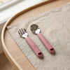 Cubkins raspberry pink silicone-handled stainless steel toddler fork and spoon set on a linen napkin on a wooden tray