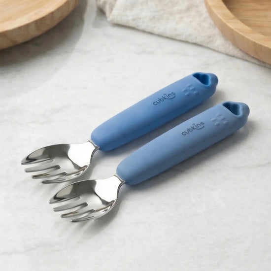 Two Cubkins blue silicone-handle stainless steel baby forks on marble surface — ergonomic grip with textured finger rest for toddler self-feeding