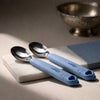 Two Cubkins blue silicone-handle stainless steel baby spoons resting on marble tile with traditional silver bowl in background — Indian kitchen lifestyle