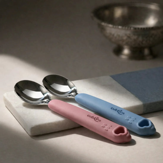 Cubkins stainless steel baby spoon set showing both blue and raspberry silicone handles on marble tile — Indian-inspired lifestyle shot with silver bowl