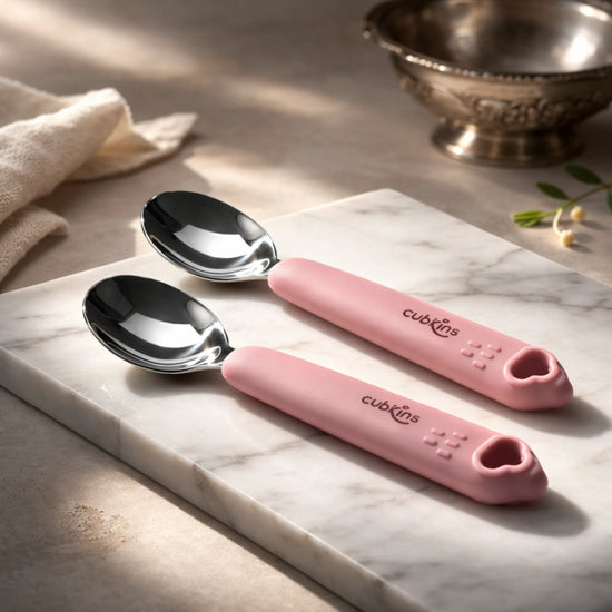 Two Cubkins raspberry silicone-handle stainless steel baby spoons on marble surface with traditional silver bowl — soft grip handle for infant self-feeding