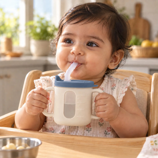 Indian baby in a high chair holding the Cubkins blue olive straw training cup by both handles and drinking water through the straw