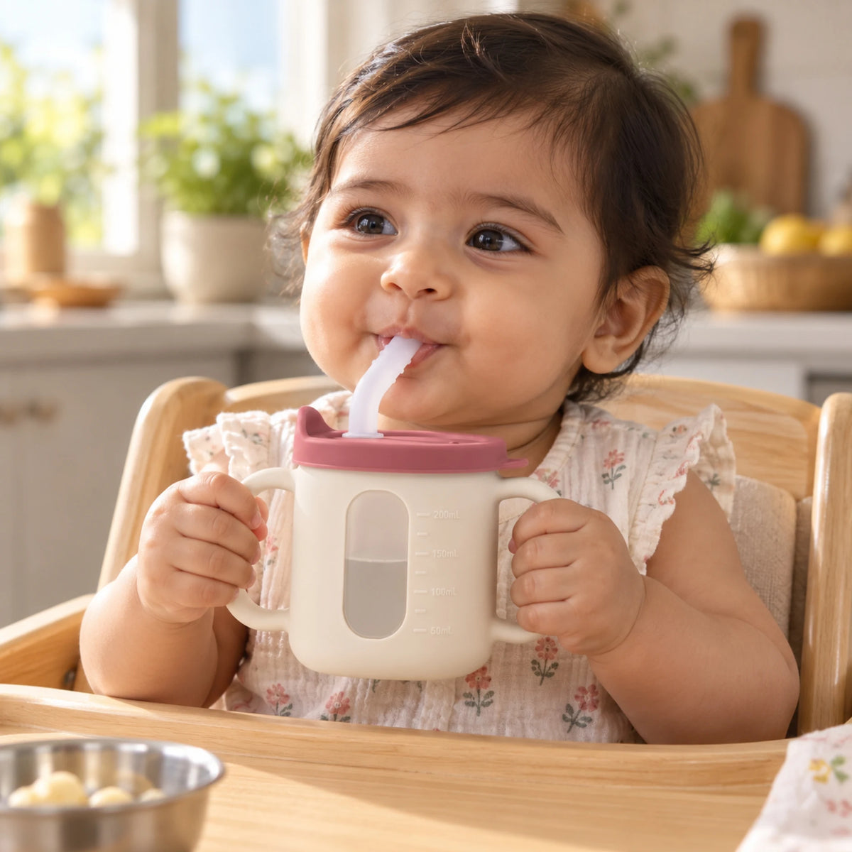 Indian baby in a high chair holding the Cubkins raspberry straw training cup by both handles and drinking water through the straw