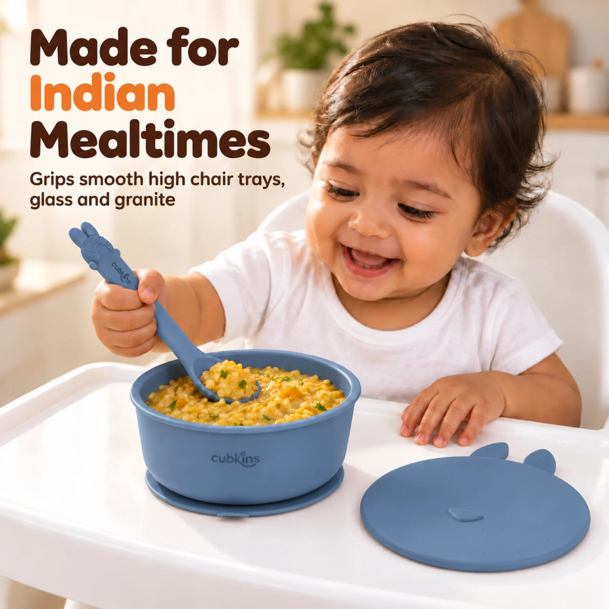 Indian baby in a high chair eating khichdi with the Cubkins Smart-Bend spoon from the blue olive silicone suction bowl stuck to the tray, its bunny lid beside it