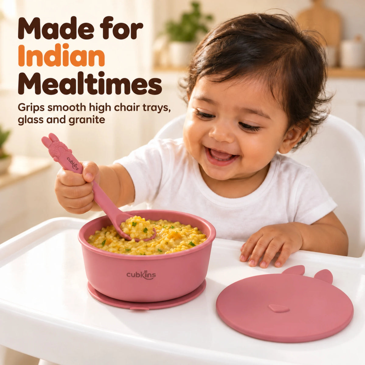 Indian baby in a high chair eating khichdi with the Cubkins Smart-Bend spoon from the raspberry silicone suction bowl stuck to the tray, its bunny lid beside it