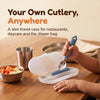 Toddler lifting the Cubkins blue olive steel spoon out of its open travel case at a restaurant table beside a steel katori of fruit