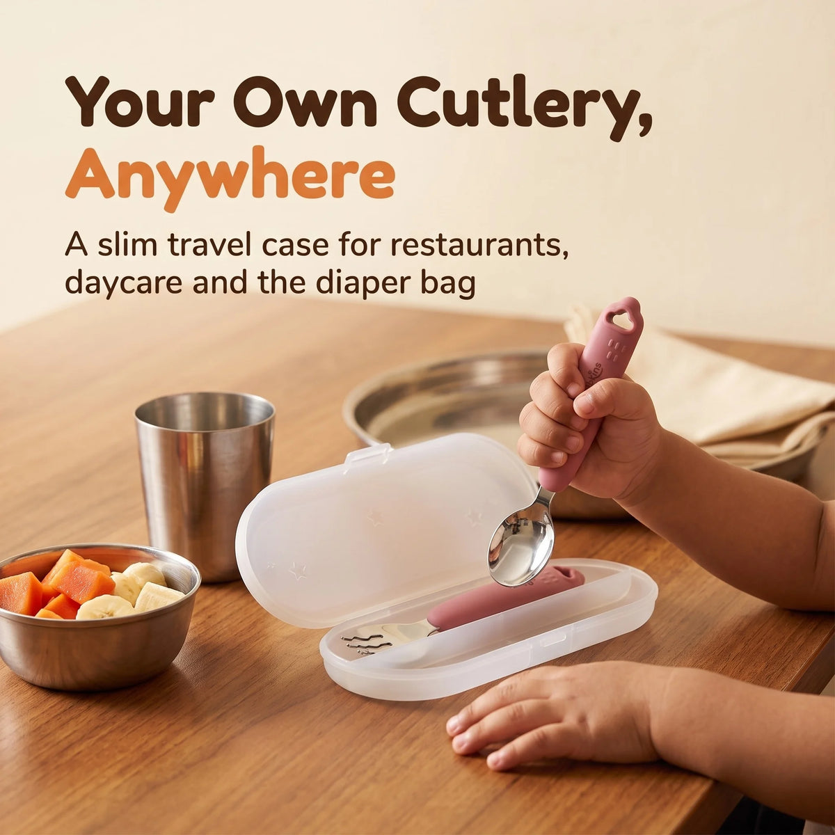 Toddler lifting the Cubkins raspberry steel spoon out of its open travel case at a restaurant table beside a steel katori of fruit