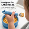 Toddler's hand holding a Cubkins Blue Olive fork with macaroni on the tines above a matching blue silicone bowl, with the text "Designed for Little Hands – Short, weighted handle improves motor control"