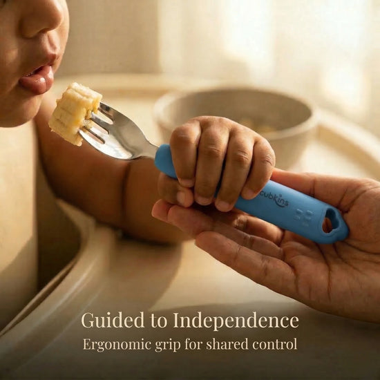Close-up of a baby and parent's hands both holding a Cubkins Blue Olive toddler fork as the baby brings a piece of banana to their mouth, with the caption "Guided to Independence – Ergonomic grip for shared control"