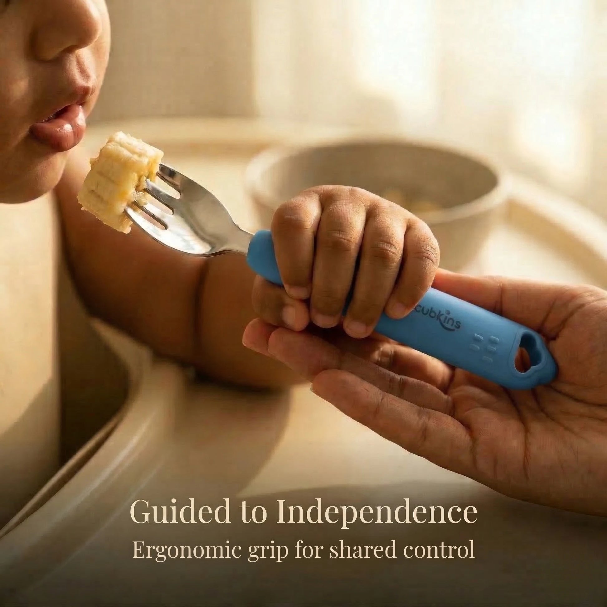 Close-up of a baby and parent's hands both holding a Cubkins Blue Olive toddler fork as the baby brings a piece of banana to their mouth, with the caption "Guided to Independence – Ergonomic grip for shared control"