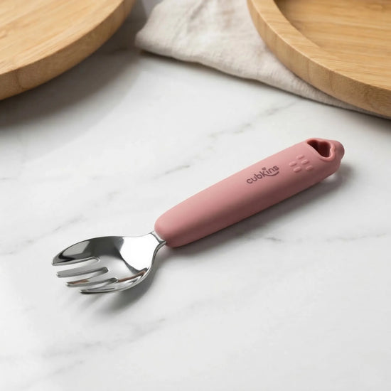 Cubkins stainless steel toddler fork in Raspberry with food-grade silicone handle, placed on a marble surface with wooden plates in the background